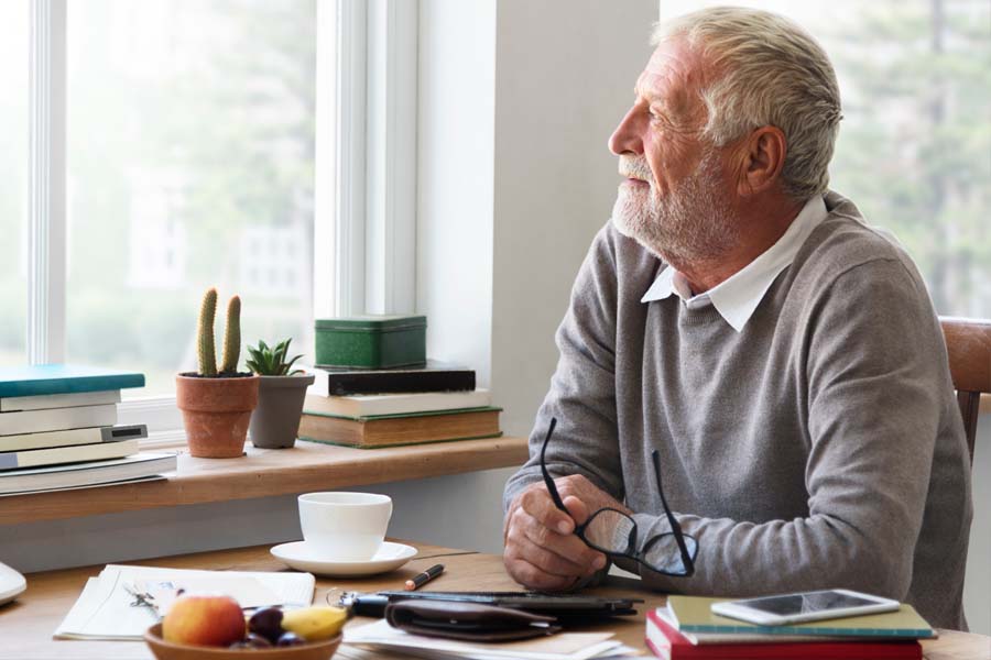 An older adult sits at a desk near a window holding glasses and looking thoughtfully toward natural light.