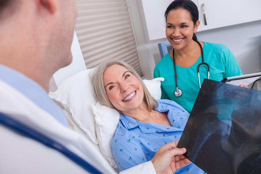 Smiling elderly woman lying on hospital bed at UTMB Health as male doctor shows her an X-ray of her hip, female nurse stands nearby