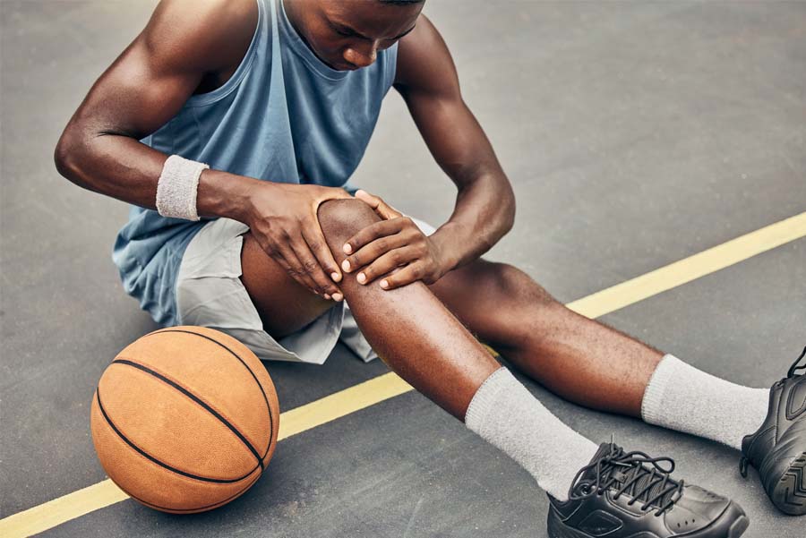 Male basketball player sitting on ground outdoors, holding his knee, after a basketball knee injury