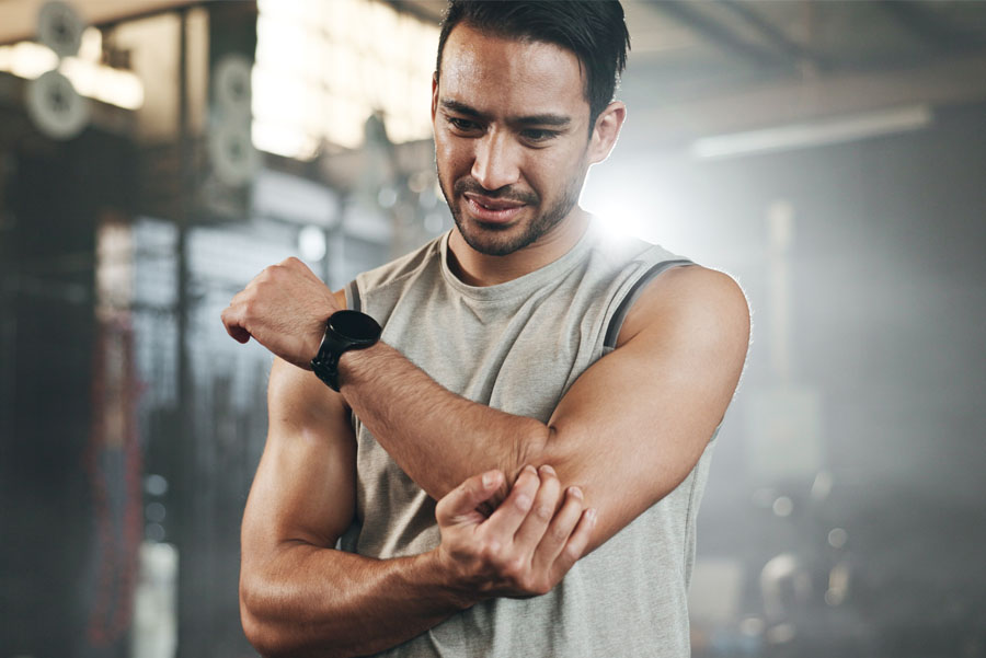 Man in gym with look of worry on his face experiencing joint problem, discomfort, or arthritis, holding his elbow