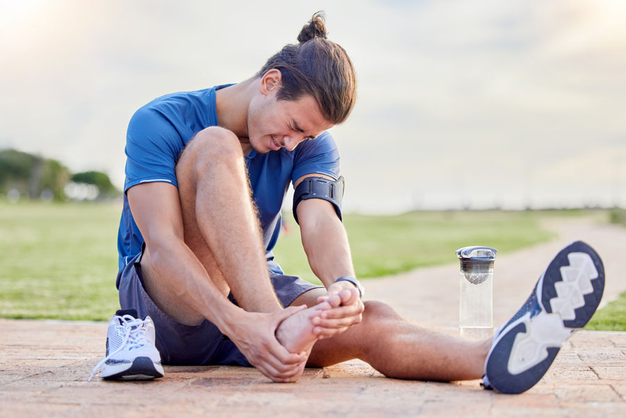 Man wincing in pain sitting on ground outdoors touching his bare foot after having removed his shoe after a running injury, water bottle next to him.