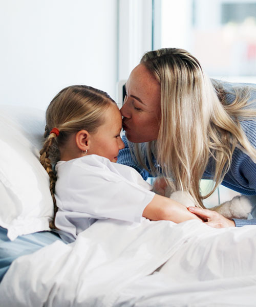 A mom kisses her daughter on the forehead while she is in a hospital bed