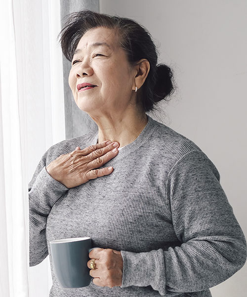 a middle aged woman holding a mug and pressing her throat with the other hand