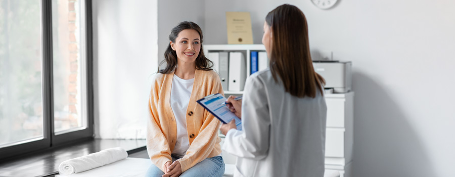 Smiling, seated female patient speaking to standing female health professional, with back to the camera, holding a clipboard