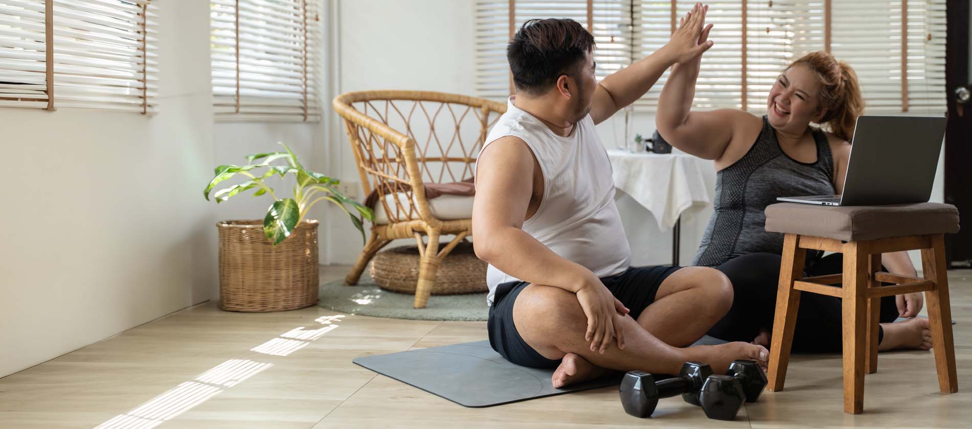Couple high fiving each other while seated on floor, about to do a workout from laptop, dumbbells nearby
