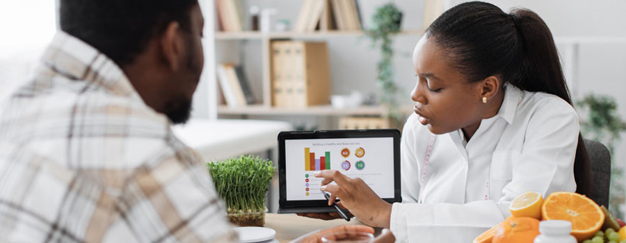 Over-the-shoulder view of a female dietician analyzing a male patient’s nutrition data on digital tablet in consulting room for weight loss treatment