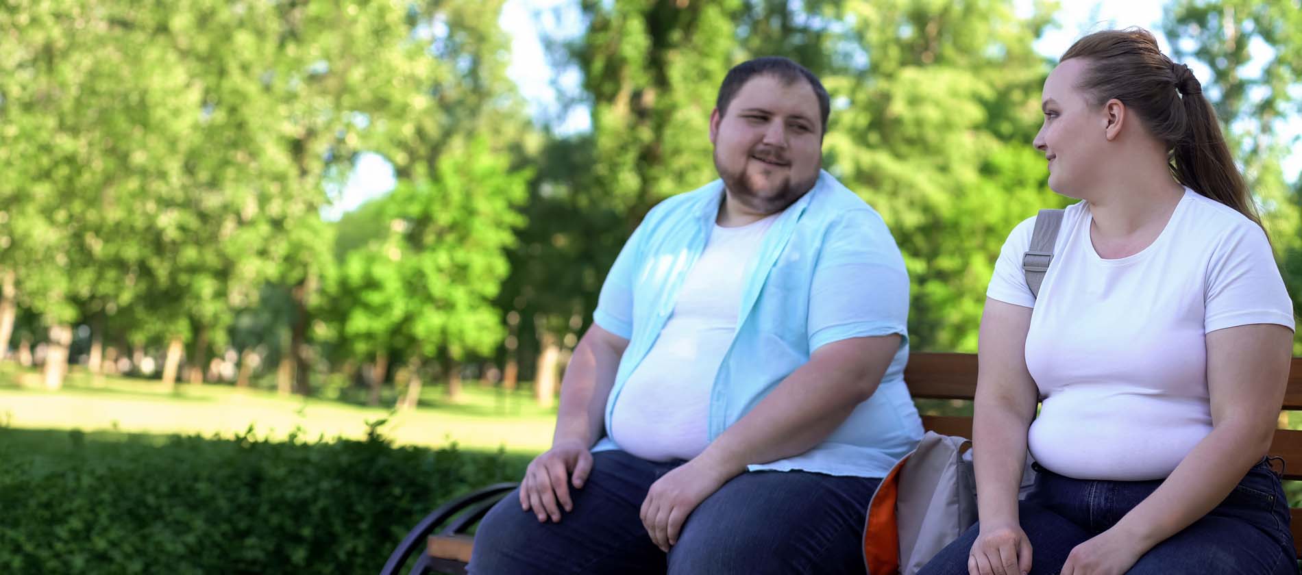 Overweight man and overweight woman looking at each other while sitting on a bench outdoors