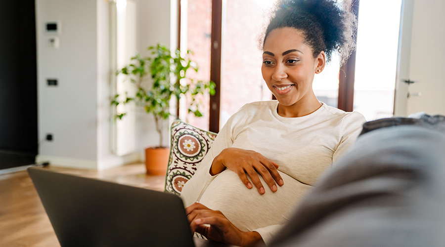 A pregnant woman smiles while using a laptop at home to learn about prenatal care from UTMB Health.