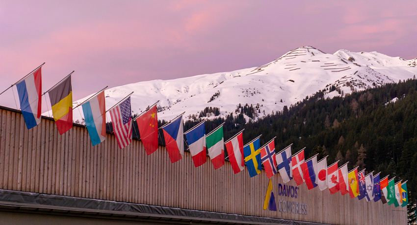 Row of international flags lining the Davos Congress Center with snow-covered mountains and a pink sky in the background.