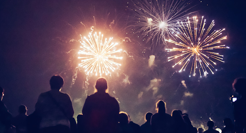 Silhouetted crowd watching multiple fireworks bursts in a night sky. Bright explosions in gold and white illuminate smoke above the onlookers.