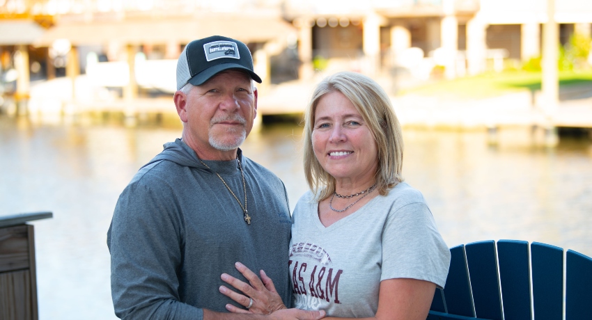 A middle-aged couple stands together outdoors near the water, smiling at the camera. The man wears a baseball cap and gray long-sleeve shirt, while the woman wears a Texas A&M T-shirt and rests her hand on his chest.