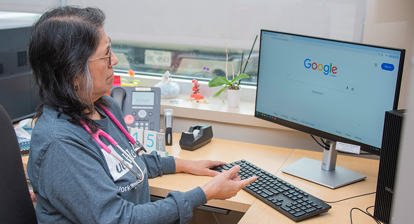 Person seated at a desk using a keyboard and looking at a computer monitor displaying the Google homepage. Office items, including a telephone, calendar, and potted plant, are visible on the desk and windowsill.