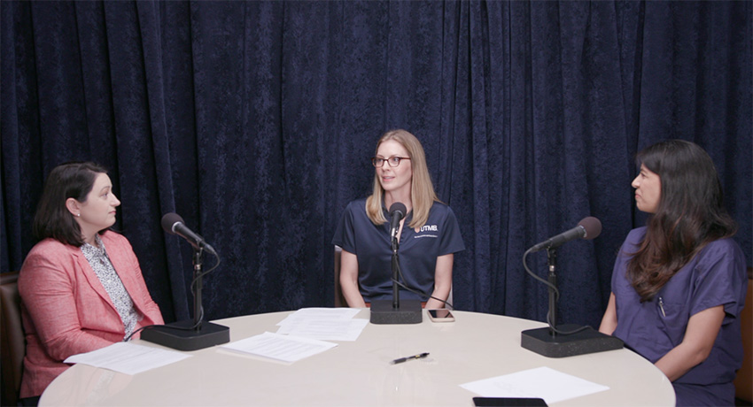 Three people sit around a round table with microphones and papers, participating in a recorded discussion in front of a dark curtain backdrop.