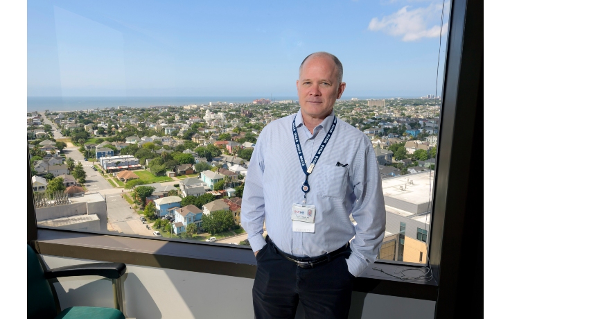 A man wearing a light blue button-down shirt and identification badge stands in front of a large window overlooking a coastal cityscape. Residential neighborhoods, buildings and the Gulf shoreline are visible in the background under a clear blue sky.