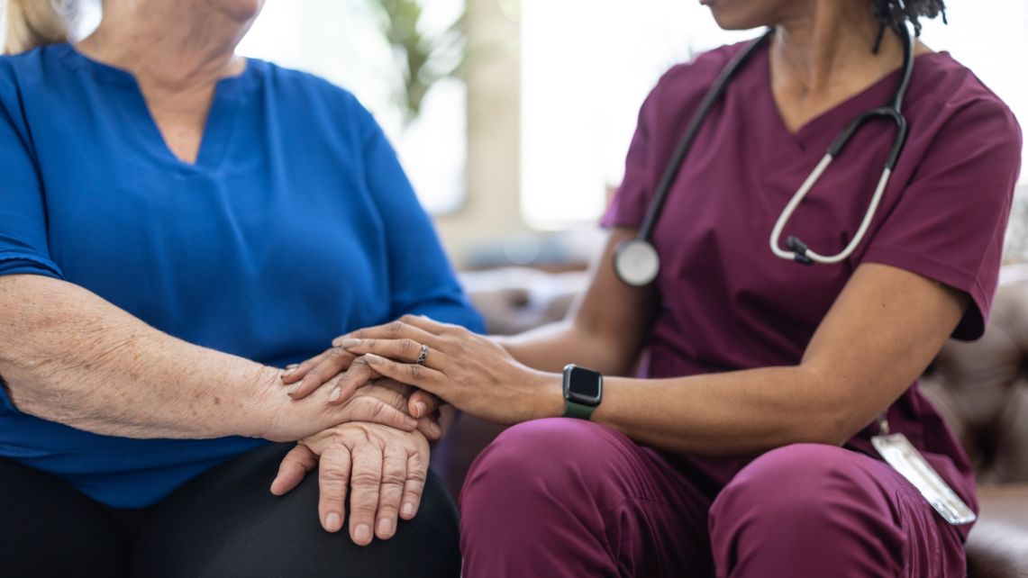 A health care worker in scrubs rests a hand on the hands of another person during a seated conversation.