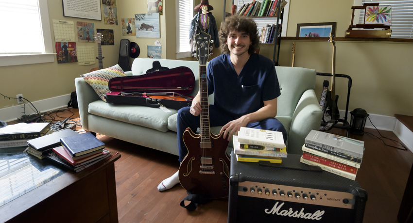 Aidan Carroll, a third-year medical student, sits in a living room holding an electric guitar. Books are stacked on a coffee table, couch and guitar amplifier around him, with an open guitar case and additional instruments in the background.