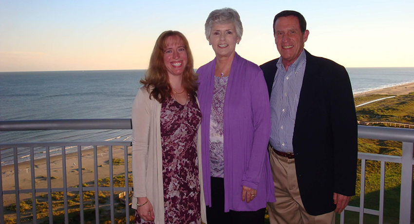 A retired couple and their adult daughter wearing dressy attire pose on a balcony in front of a beach at sunset.