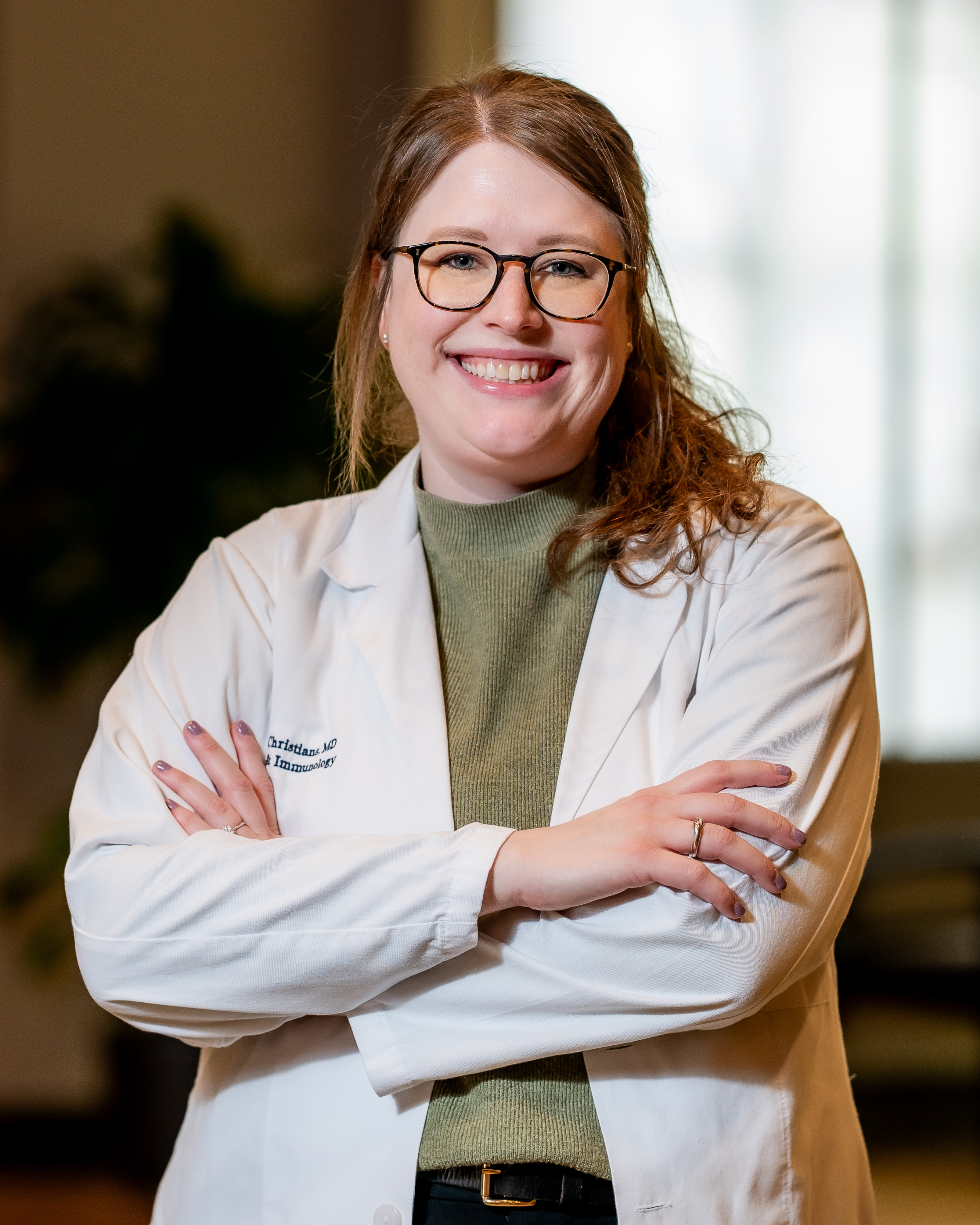 Healthcare professional in a white coat with a UTMB Health logo, wearing glasses and standing with arms crossed indoors against a softly blurred background.