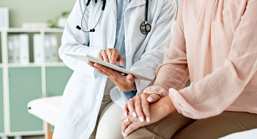 Clinician wearing a white coat and stethoscope holds a tablet while seated next to a patient, with both focused on the screen during a consultation.