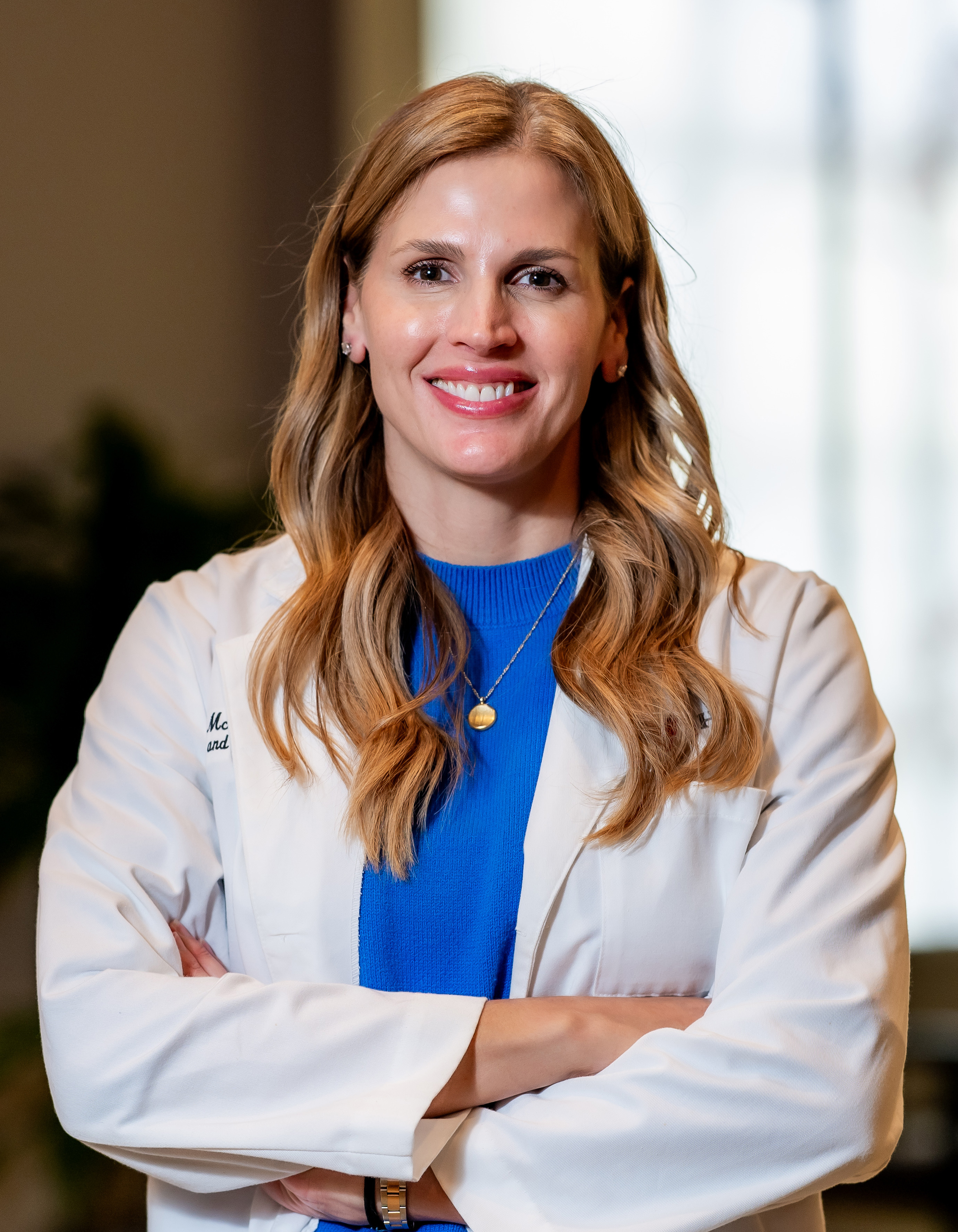 A person in a white lab coat with arms crossed stands indoors in front of a bright window.