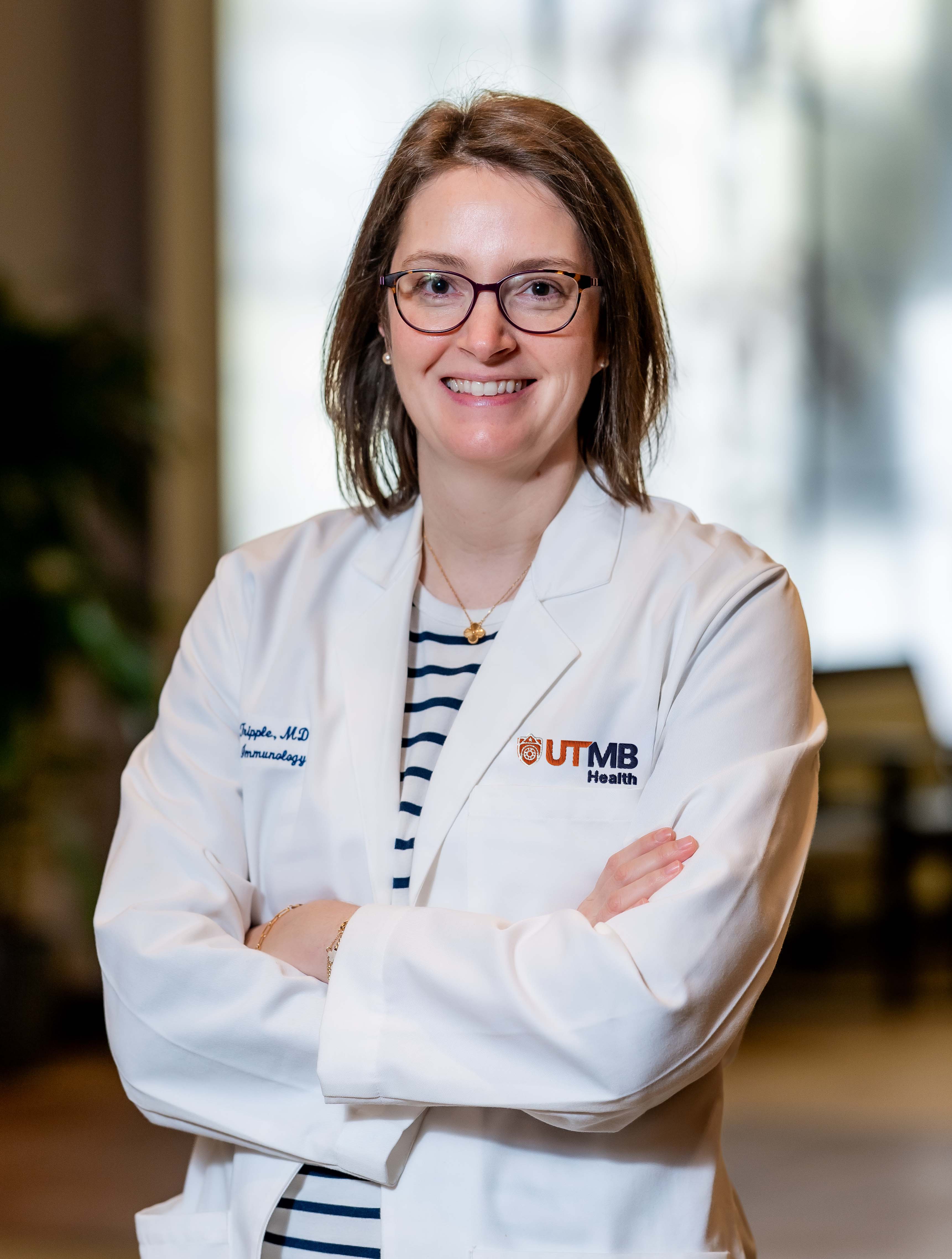 Clinician in UTMB Health lab coat standing with arms crossed A person wearing a UTMB Health white lab coat stands indoors with arms crossed, shown from the waist up against a softly blurred background.