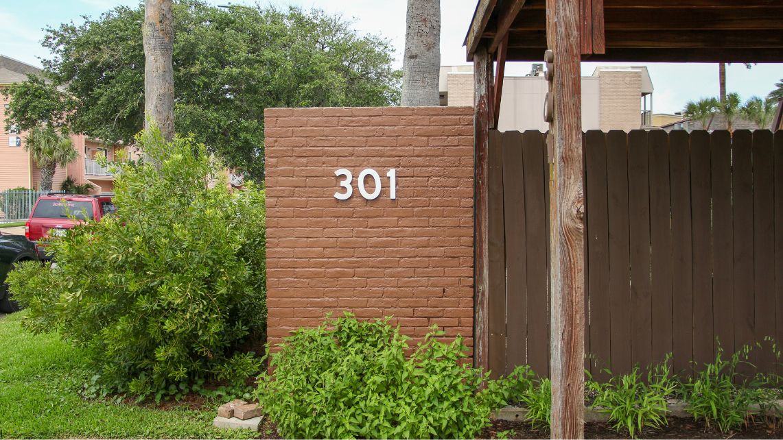 Exterior view of David’s House in Galveston, showing a brick wall marked with the number “301,” a wooden fence, greenery, and nearby residential buildings, representing a community resource supporting individuals experiencing homelessness
