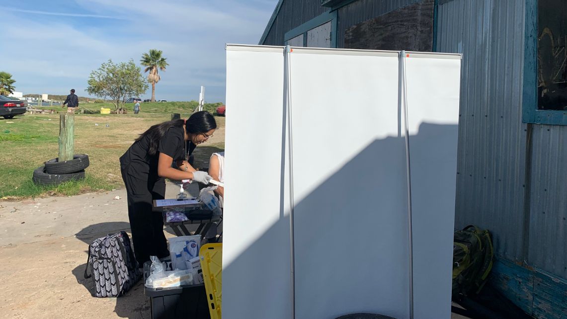 A nurse in black scrubs talks to a patient sitting behind a screen at an improvised dockside clinic