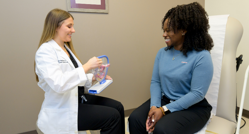 A healthcare provider in a white coat uses a pelvic model to explain anatomy to a smiling patient seated on an exam table in a clinical exam room.