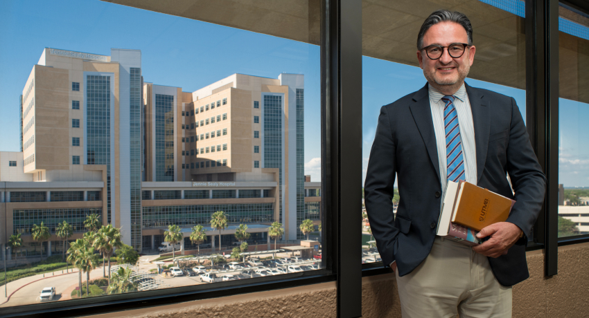 A man in a suit with short dark hair and black glasses stands indoors by a window holding books, with the Jennie Sealy Hospital building visible outside.