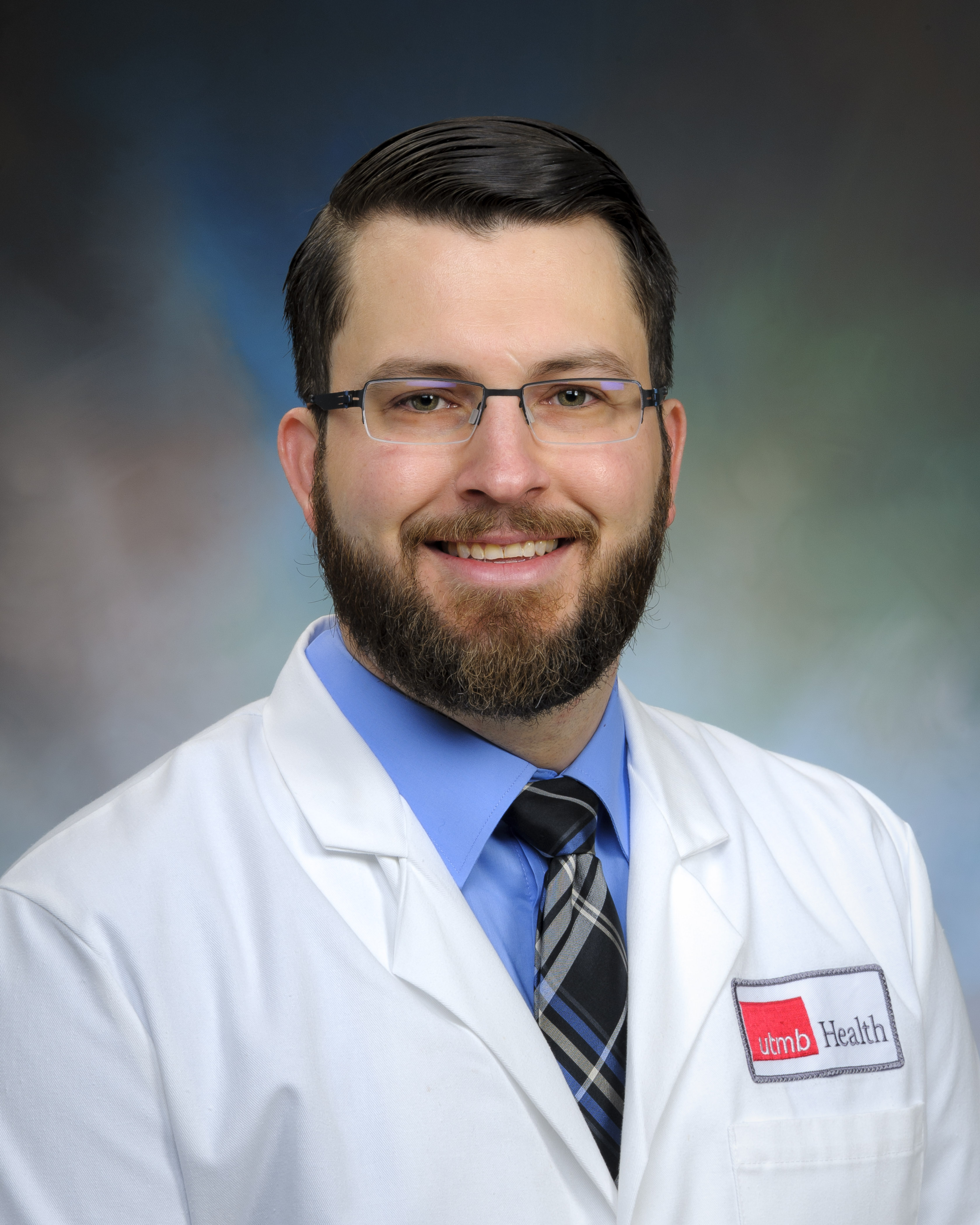 Person wearing a white lab coat with a UTMB Health logo patch, blue shirt, and patterned tie, posed against a neutral studio background.