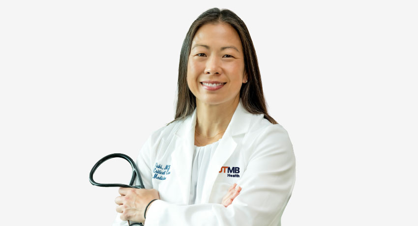 A woman stands smiling in a white UTMB Health lab coat while holding a stethoscope with her arms crossed.