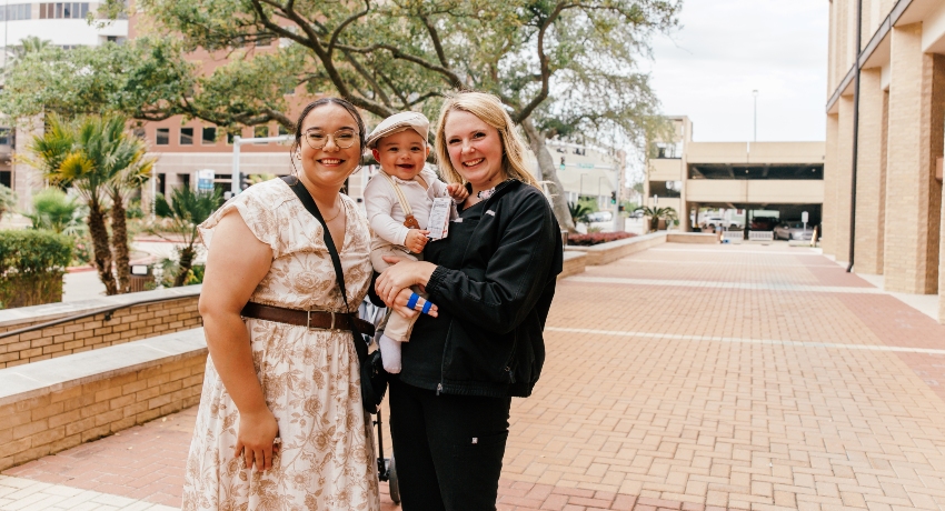 “Three people stand on a brick walkway outside a campus building. One adult holds a baby wearing a light-colored outfit and cap. Another adult stands nearby in a light patterned dress and glasses. Trees, planters, and buildings appear in the background.”