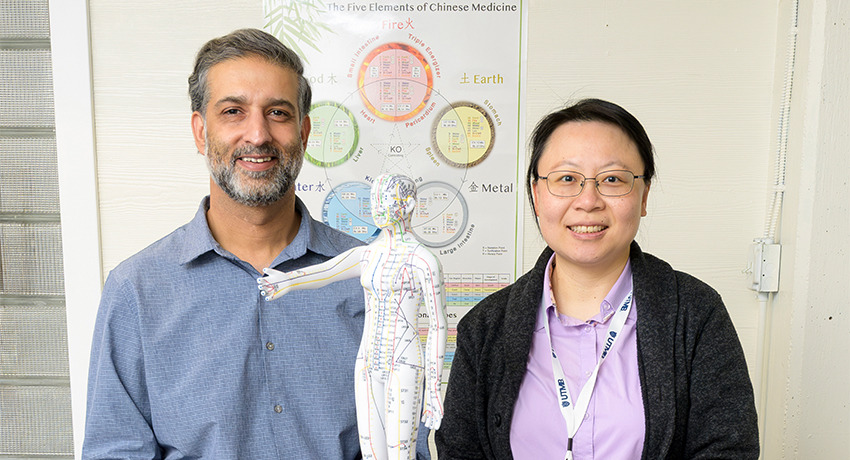 Two people stand behind a table, smiling at the camera, with a detailed acupuncture model placed between them. Behind them is a wall poster showing diagrams related to traditional Chinese medicine and the body’s meridian system