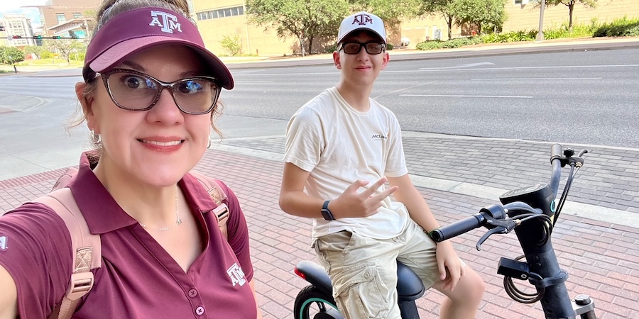 Two people outdoors on a sidewalk, one seated on an electric bike and the other standing beside it, with a street and buildings in the background