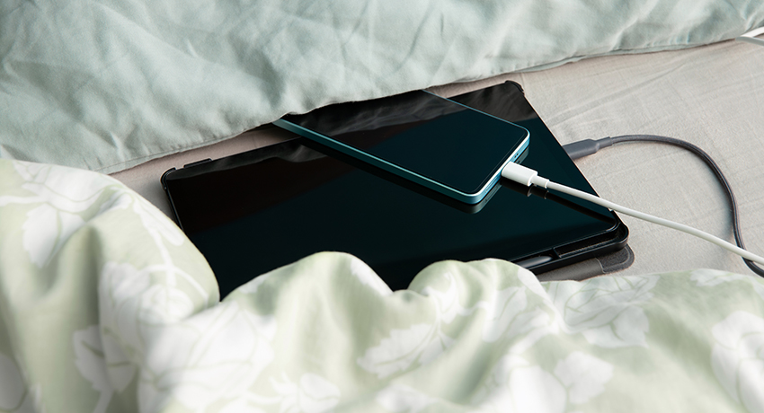 A tablet and a cell phone, both with charging cords attached, rest on a bed next to a comforter and a pillow.