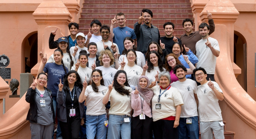About two dozen diverse medical students stand on outdoor steps, smiling and raising one finger to celebrate being first in their families to attend medical school, wearing matching light shirts in front of a brick staircase.