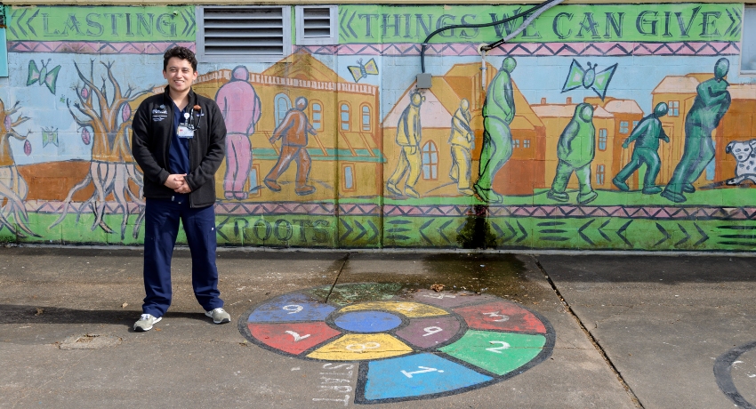 A person in medical scrubs and a jacket stands smiling on a sidewalk in front of a colorful mural depicting figures, buildings, and symbolic scenes. Painted on the ground in front of them is a circular, multicolored design divided into numbered sections.