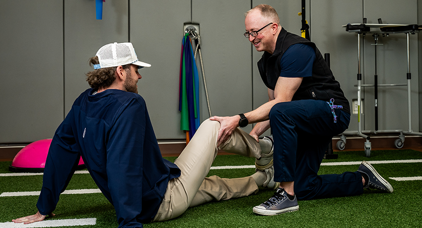 A person kneels on indoor turf and assists another seated person with a leg movement exercise. Resistance bands and rehabilitation equipment are visible in the background.