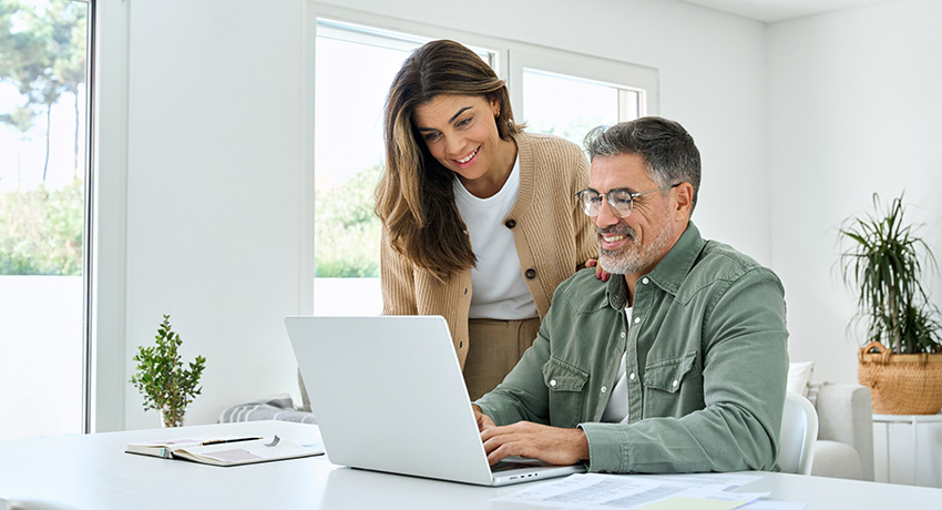 Two people review information on a laptop at a table in a bright room with papers and a notebook nearby.