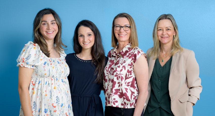 Four women in business casual clothing stand close together in front of a blue background.