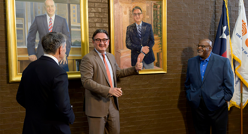 Three people stand in front of two framed portraits on a brick wall, with one person gesturing toward the artwork. Flags with partial insignia are positioned to the right.