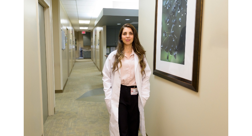 A person wearing a white lab coat stands in a well-lit medical office hallway. Framed artwork hangs on the wall beside the individual, and exam room doors and clinical workspaces are visible in the background.