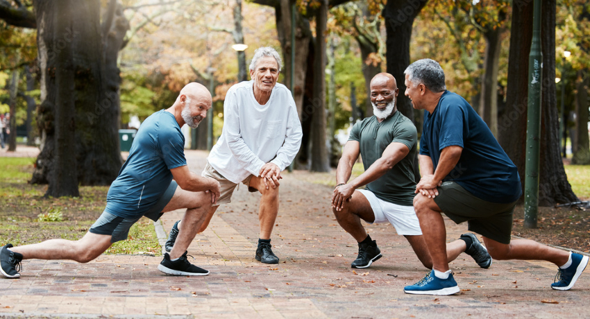 Four adults stretching in a park, each performing a forward lunge on a paved path with trees and fall foliage in the background.