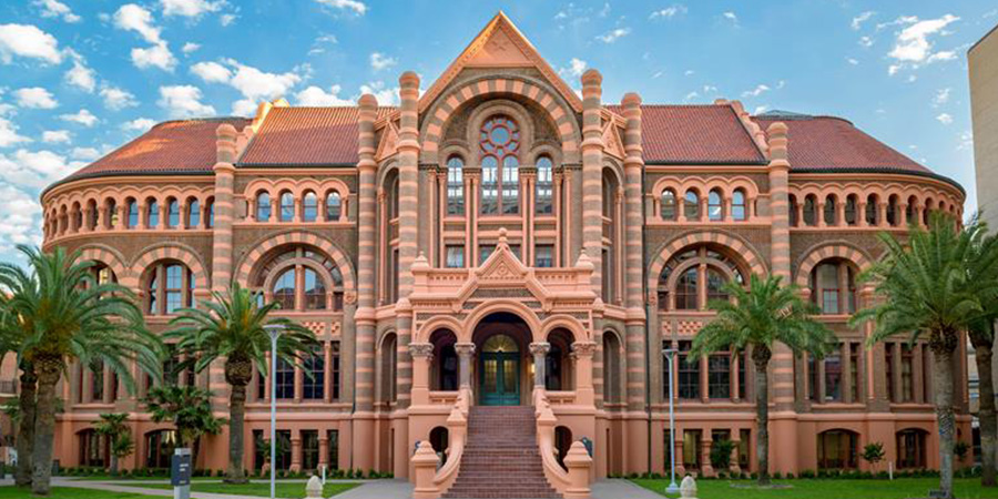 Frontal exterior view of UTMB Old Red building, featuring red brick architecture, arched windows, and palm trees lining the entrance under a blue sky