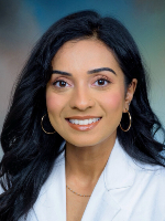 Professional headshot shows a woman with dark hair wearing a white coat against a blue-gray background, facing the camera.