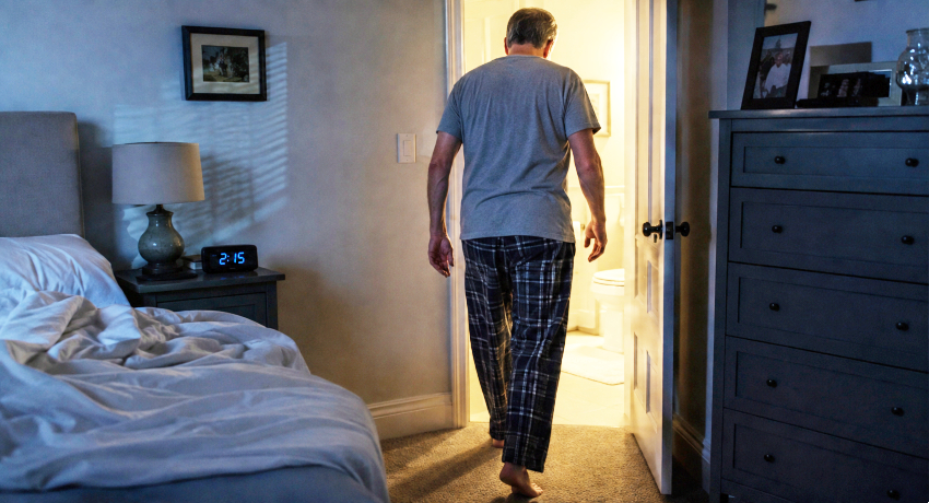 An older man walks from his bedroom to a brightly lit bathroom during the night, illustrating nighttime bathroom trips associated with an enlarged prostate.