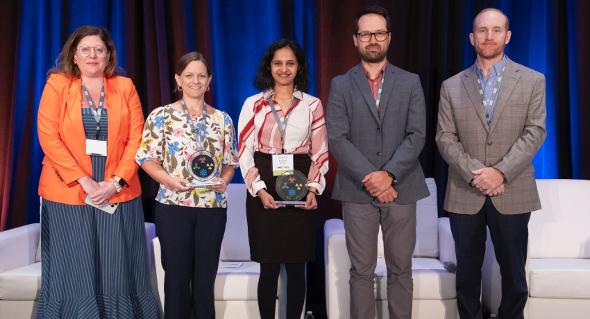Five adults stand on a stage with blue and red lighting, with two individuals holding clear glass awards.