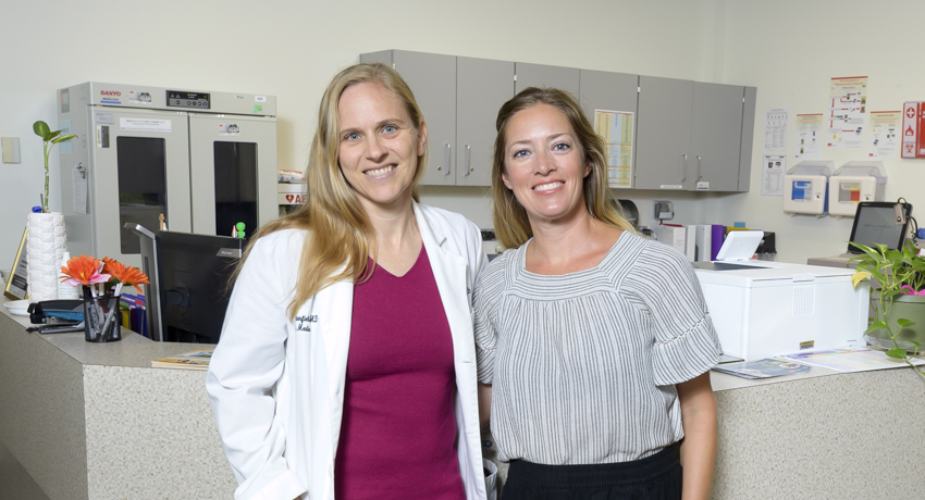 Dr. Laura Porterfield and Christen Walcher, MPAff, stand side by side in a primary care clinic at UTMB Health. The pair collaborated on research published in Annals of Family Medicine examining the paradoxes of integrating social care into primary care.