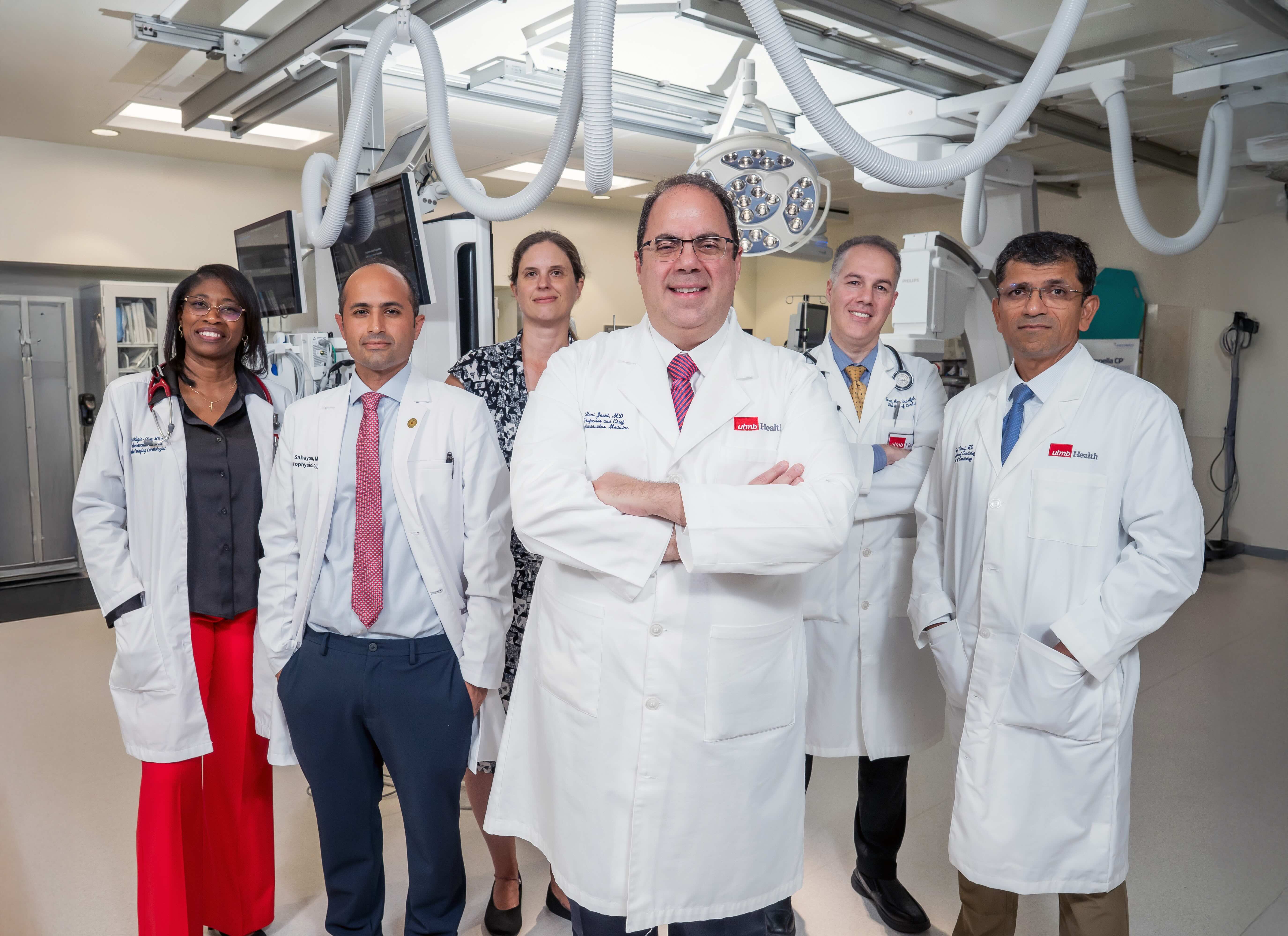 Five people wearing white lab coats stand together inside a clinical procedure room with medical equipment, overhead lights and monitors visible in the background