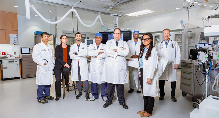 Seven men and women in UTMB white coats and one woman in black business attire stand in a procedure room.
